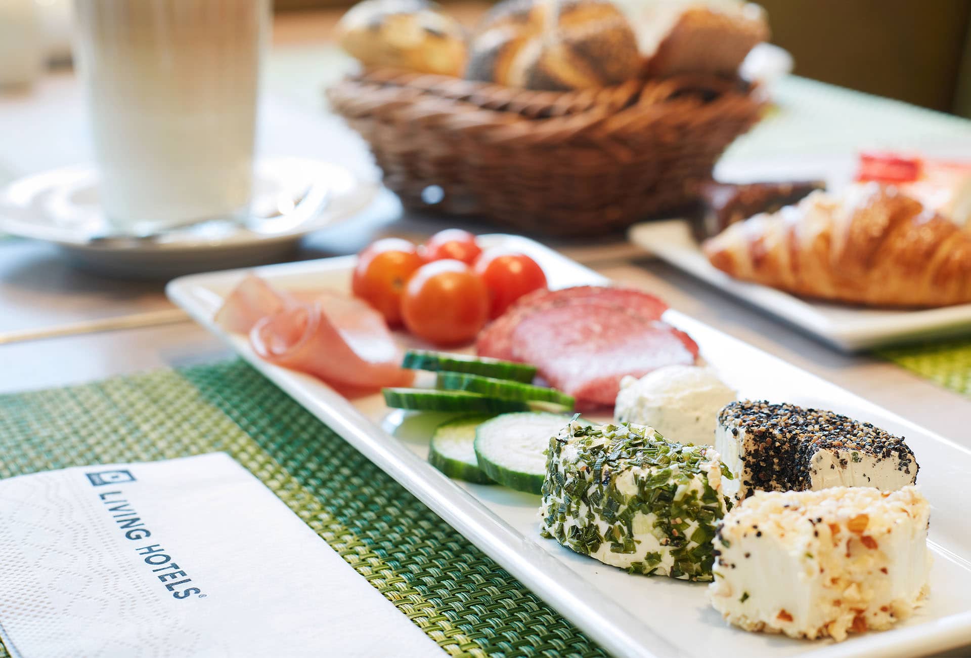Frühstücksmitarbeitende gedeckter Frühstückstisch mit einem Teller aufgereihtem Frischkäse, Gurken und Wurstaufschnitt